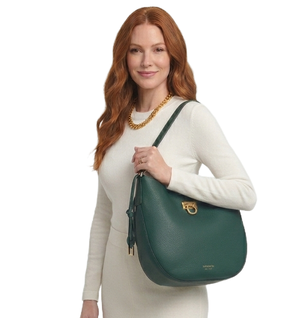 A woman with long wavy red hair wearing a white knit sweater and gold chain necklace, holding a structured emerald green pebbled leather hobo bag with gold hardware against a minimalist white studio background.